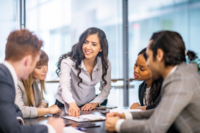 Woman sitting at a table surrounded by employees, she is gesturing at a business meeting with confidence.